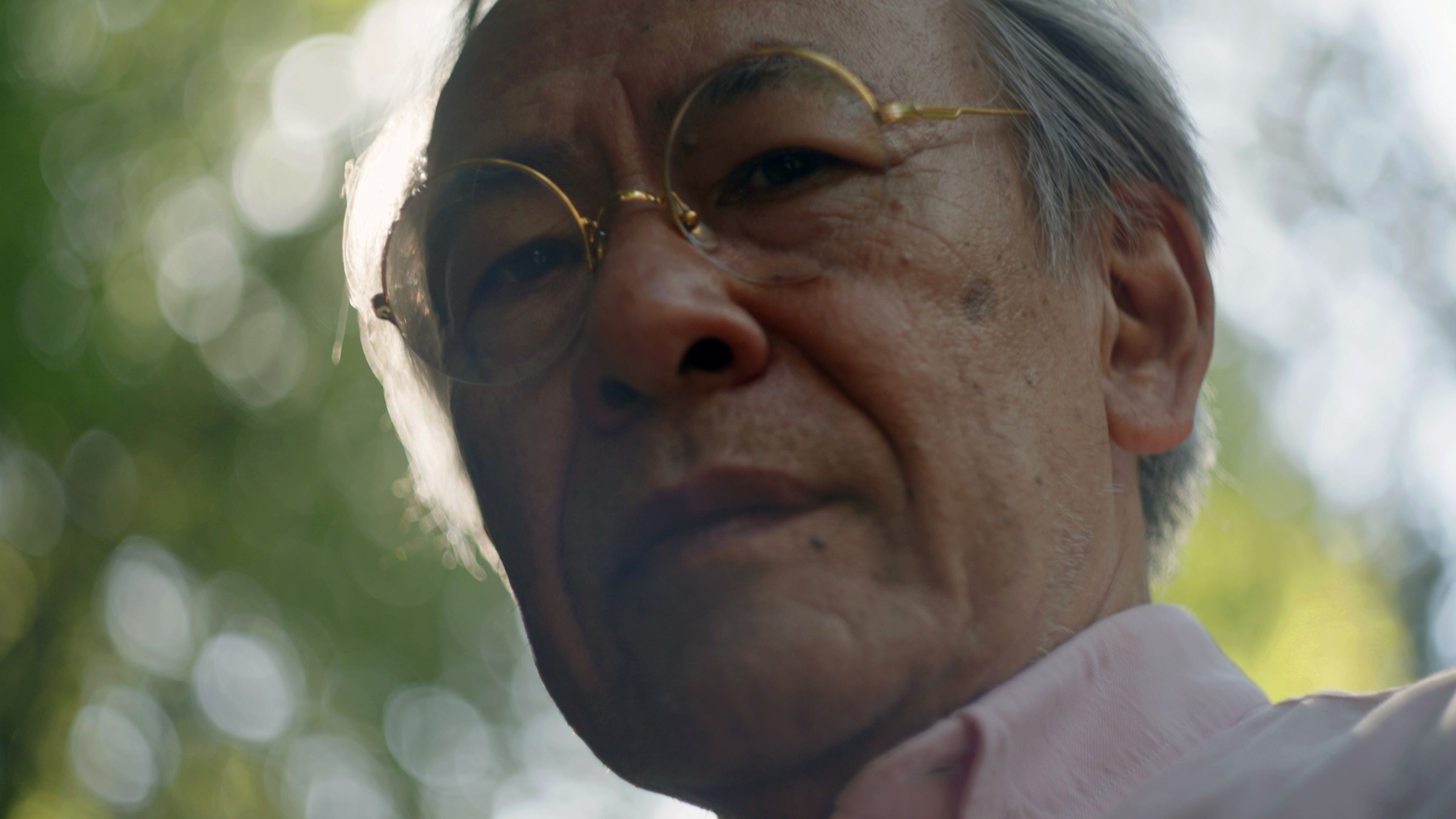 Close portrait of Seiichiro Tatsumi in round gold glasses, green trees behind him